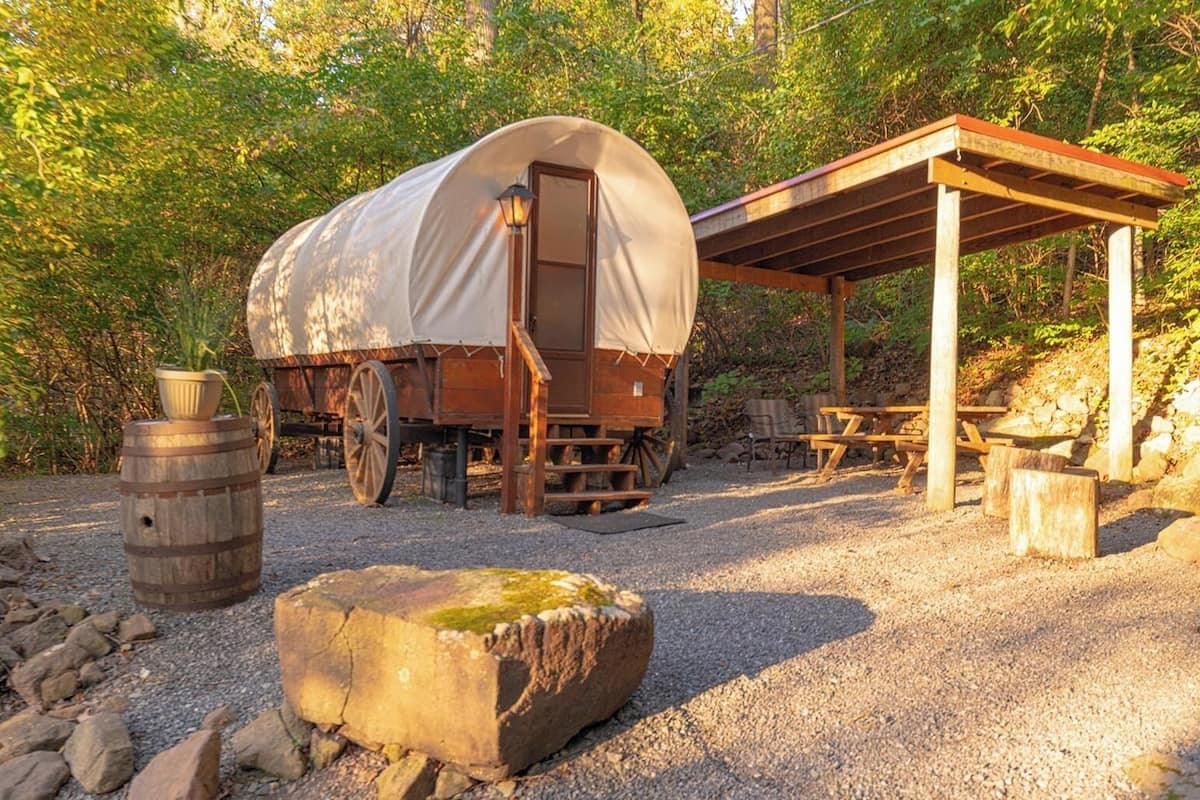 A covered wagon with a wooden structure and picnic tables in a forested area.