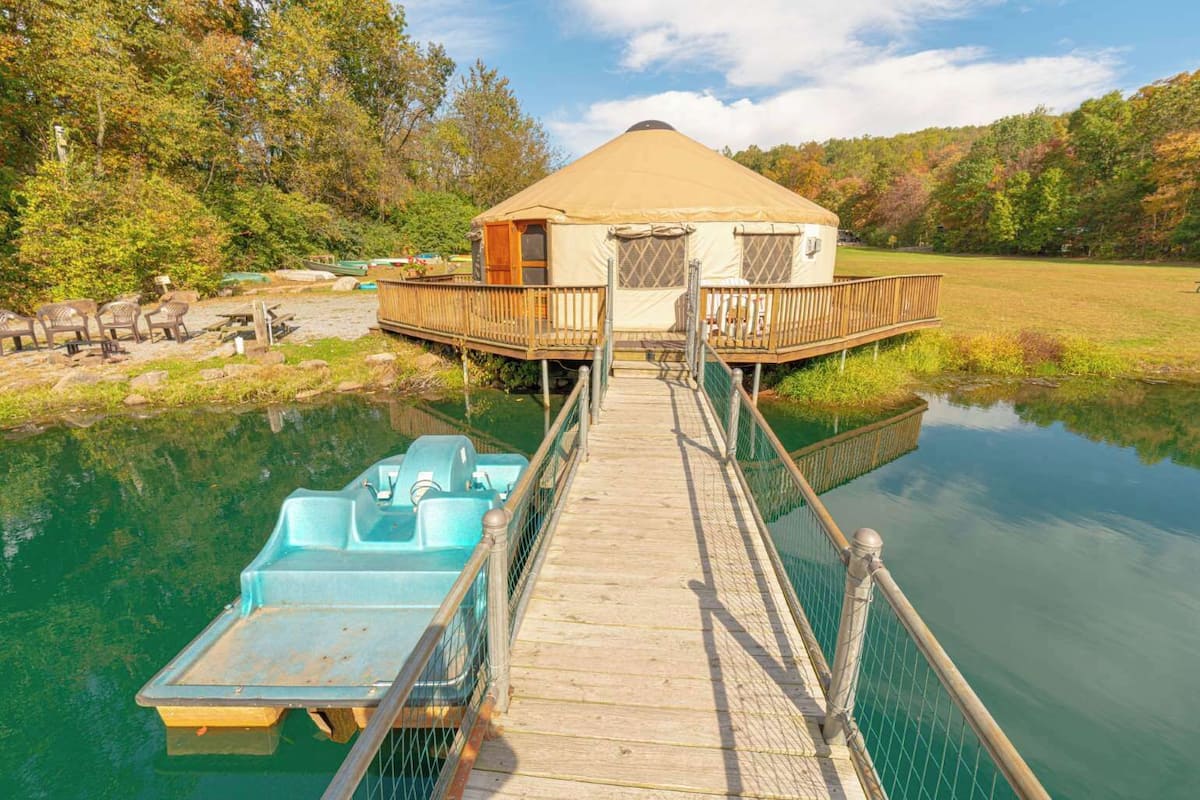 A yurt with a dock extending into a calm body of water, surrounded by trees and a grassy area.