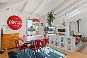 A modern living room with a dining area, featuring a red Coca-Cola sign, a large plant, and a view of the outdoors. - Paddle, Relax, Repeat on Seadrift Lagoon (Stinson Beach)