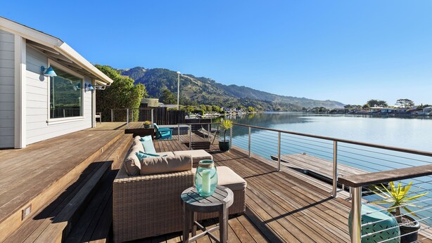 Una terraza de madera con mobiliario exterior, vista a un lago y montañas al fondo.