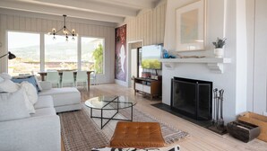 A living room with a sofa, coffee table, fireplace, and a TV. There are large windows and a view of the outdoors.