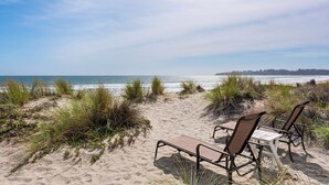 Una playa con dos sillas de descanso vacías y una mesa, rodeada de arena y césped.