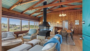 Interior acogedor de una cabaña con chimenea de piedra, mobiliario de mimbre y amplias ventanas que ofrecen vistas al exterior.