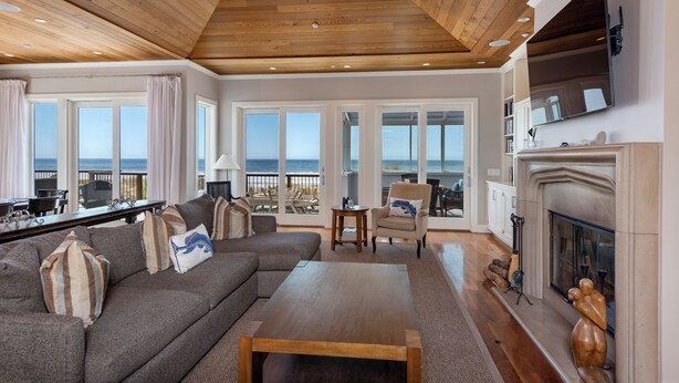 A spacious living room with a wooden ceiling, large windows, a fireplace, and a view of the ocean.