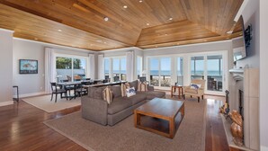 A spacious living room with a wooden ceiling, large windows offering a view of the ocean, a cozy seating area with a sofa and coffee table, and a fireplace.