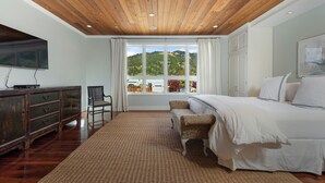 A bedroom with a wooden ceiling, a large window with a view of hills, a bed, a dresser, and a chair.