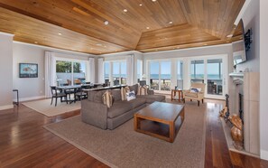 Een ruime woonkamer met houten plafond, grote ramen met uitzicht op de oceaan, een gezellige zithoek met een bank en salontafel, en een open haard.