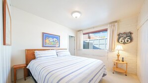 Une chambre à coucher comprenant un lit, une table de chevet, une lampe, un tableau et une fenêtre donnant sur une maison.
