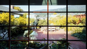 Une vue à travers une fenêtre à carreaux, dévoilant un patio avec une table et des chaises, entouré de verdure et bordé d’un chemin en briques.