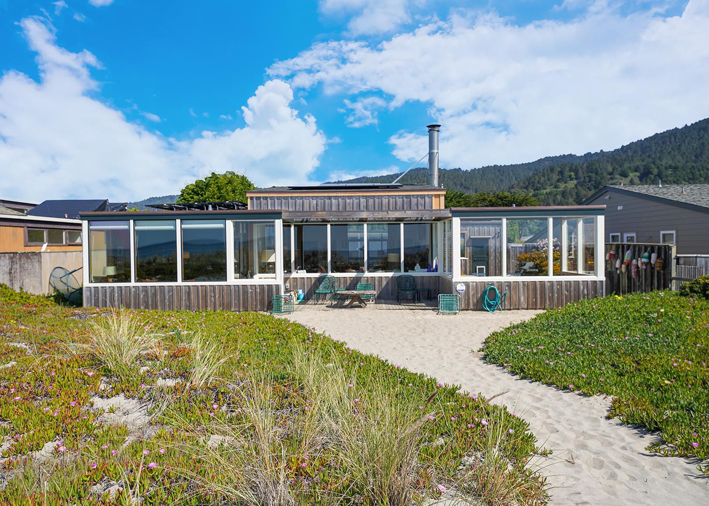 A modern house with large windows, a wooden deck, and a sandy pathway leading to the entrance.