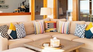A beige sectional sofa with striped and patterned cushions, a wooden coffee table with a tissue box, and a view of the ocean through large windows.
