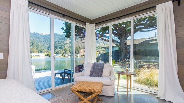 A modern room with a large sliding glass door leading to a deck with a view of a lake and hills. There is a wicker ottoman and a small round table with a lamp.