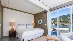 A bedroom with a large bed, a nightstand, a lamp, a wicker chair, and a view of a hot tub and hills.