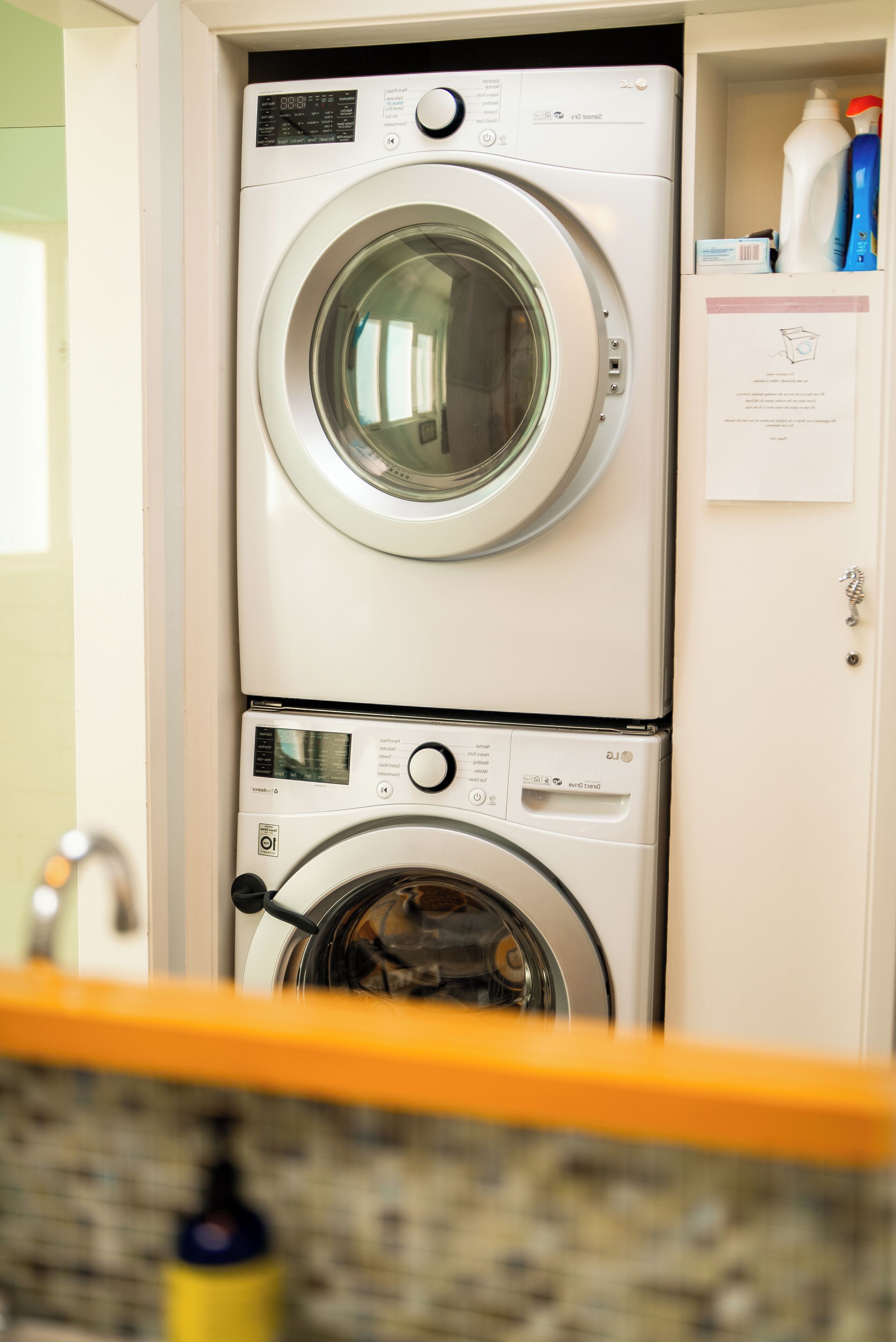 A stacked washer and dryer unit with a control panel, a yellow towel rack, and a storage cabinet with cleaning supplies.