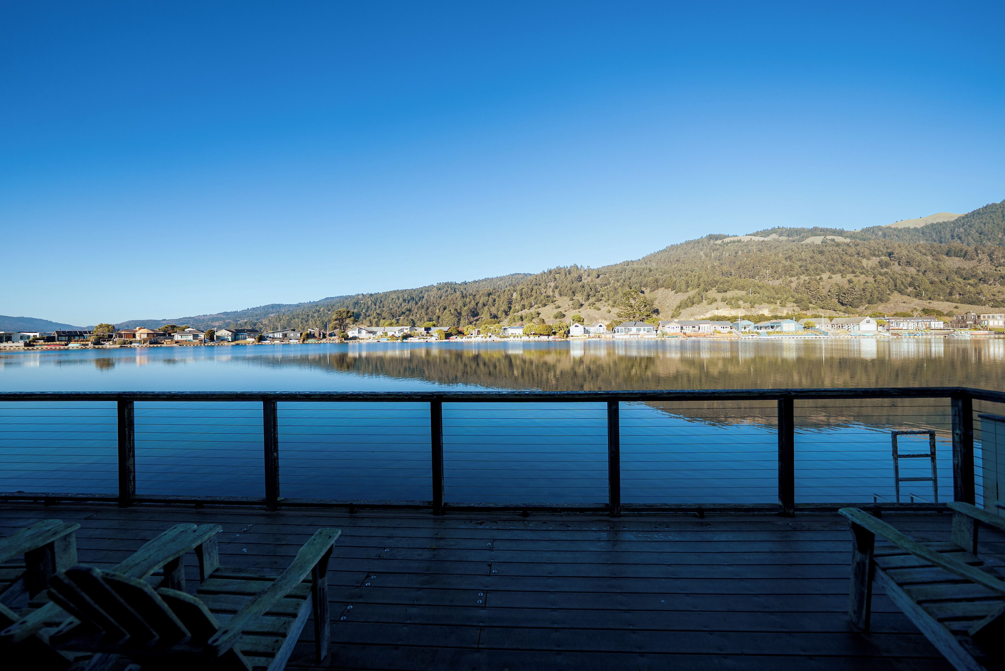 Eine Holzterrasse mit Blick auf einen stillen See, im Hintergrund eine Stadt und Hügel.
