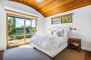 Quarto com uma cama grande, mesa de cabeceira de madeira, janela com persianas e vista para o verde lá fora.