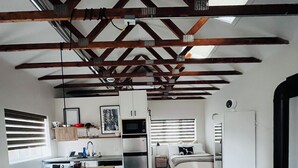Ein modernes Interieur mit freiliegenden Holzbalken, einer Küche mit Kühlschrank, einem Essbereich mit Tisch und Stühlen sowie einem Schlafzimmer mit Bett und Fensterläden.