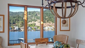A dining area with a view of a lake and hills, wooden furniture, and a chandelier. - Light and Breezy Seadrift Lagoon Home w/ Hot Tub (Stinson Beach)