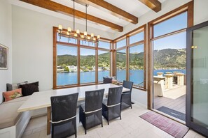 A dining area with a table and chairs, large windows with a view of a lake and hills, and a wooden ceiling.