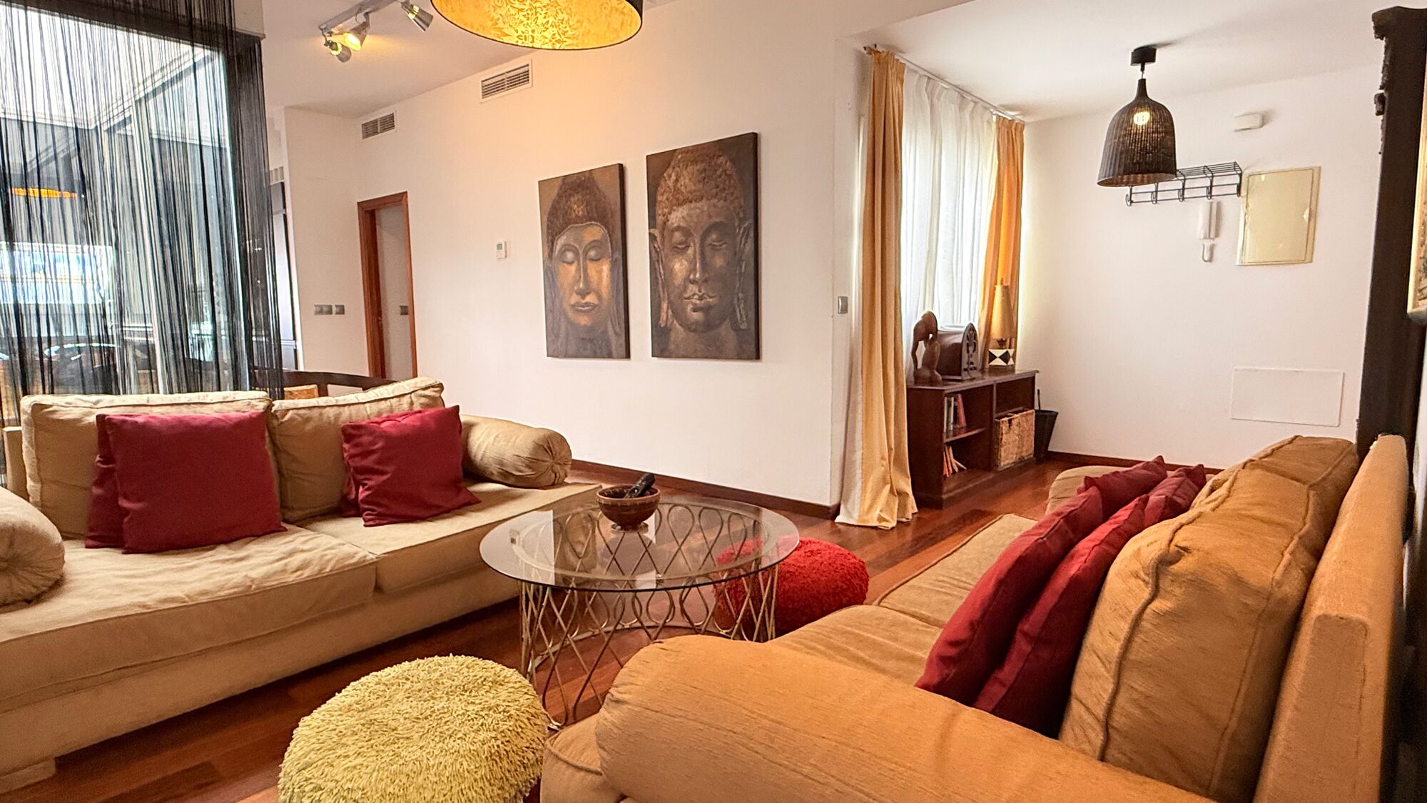 A living room with a sofa, ottoman, and a glass coffee table. There are two Buddha statues on the wall, a bookshelf, and a hanging light fixture.
