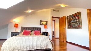 Um quarto com uma cama grande, duas luminárias de cabeceira, uma luz de teto, uma porta que dá para outro cômodo e um quadro pendurado na parede.