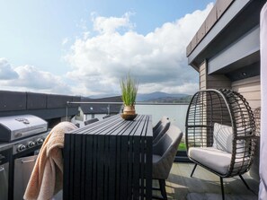 Una terraza en la azotea con una parrilla, una mesa con sillas, una planta en maceta y una silla de mimbre.