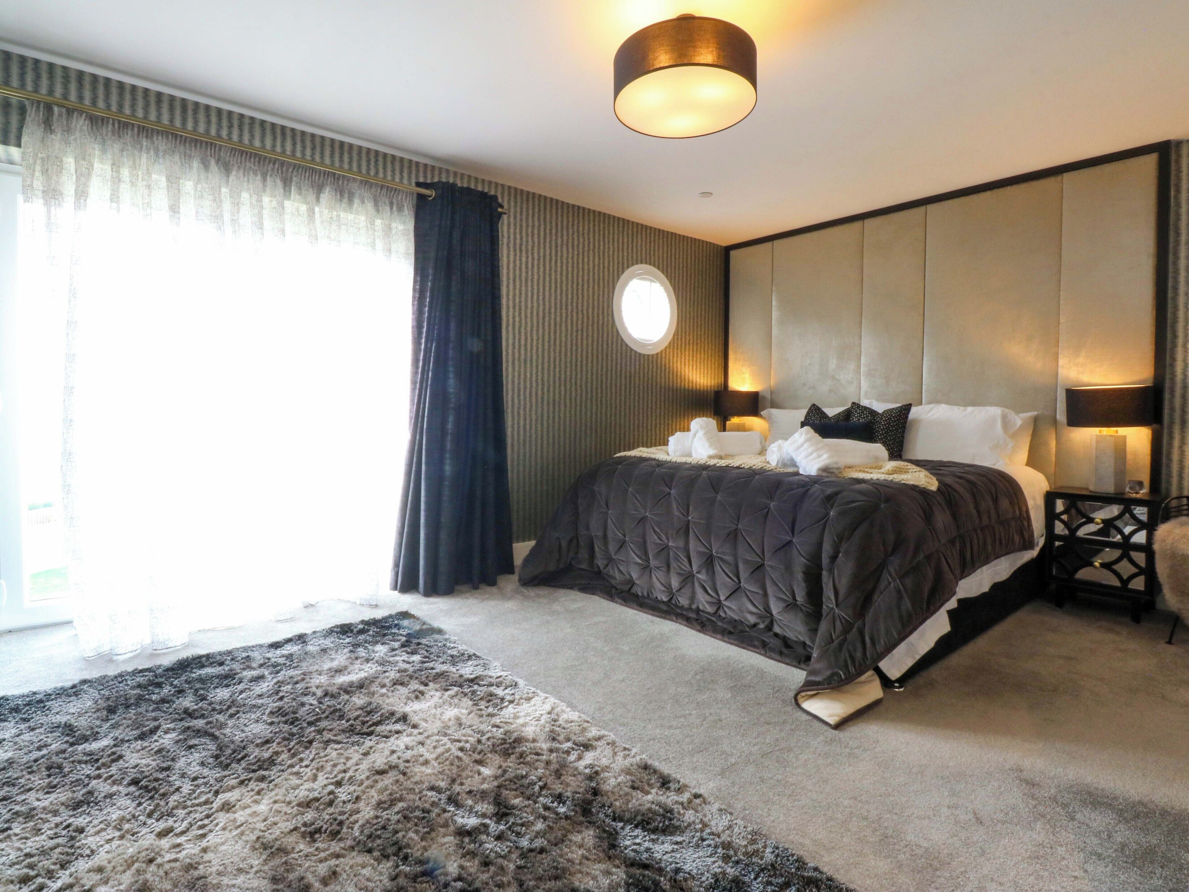A modern hotel room with a large bed, a round window, and a dark-colored headboard.
