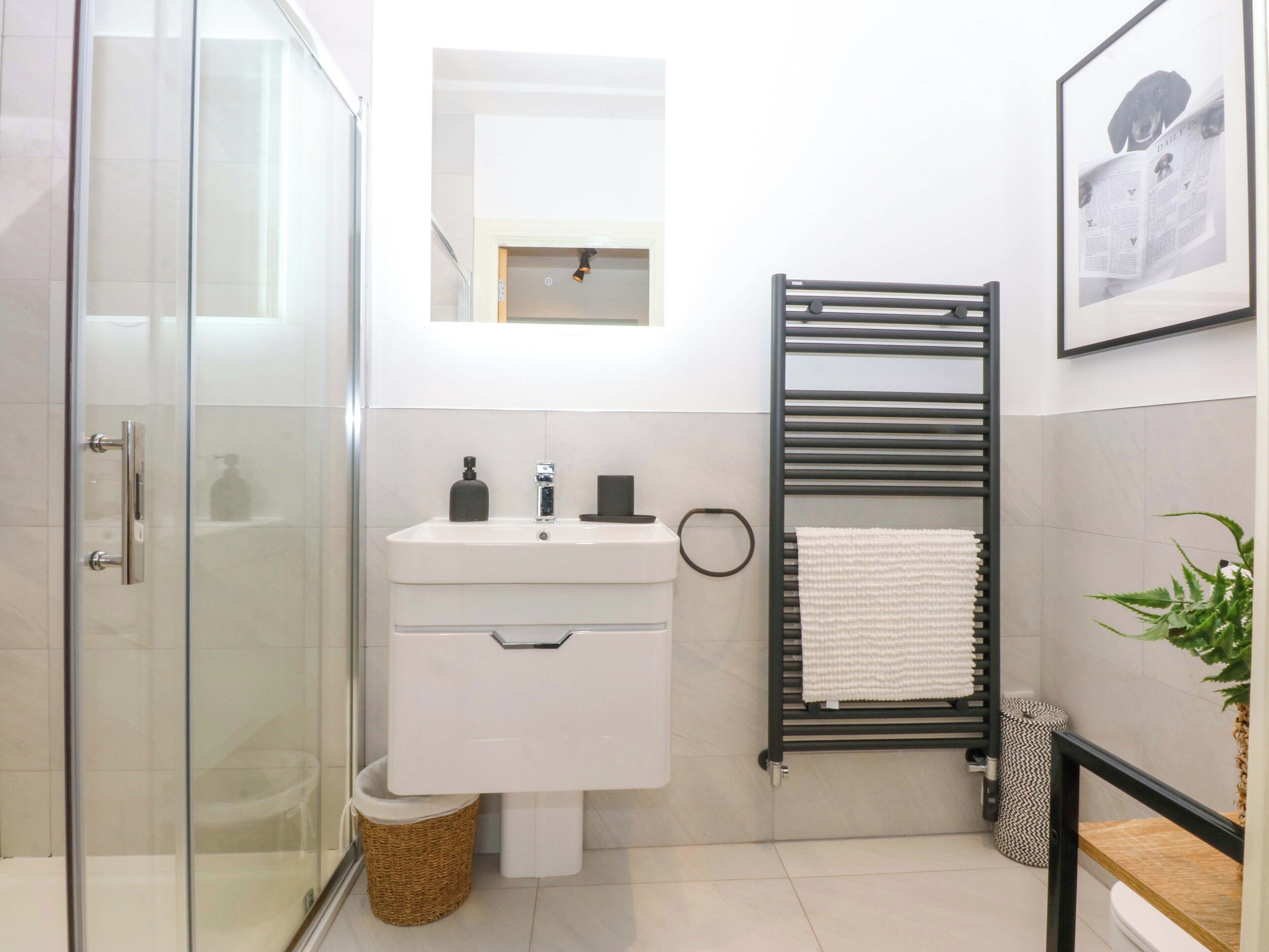 Un baño moderno con cabina de ducha de vidrio, un lavabo blanco con espejo encima, un perchero negro y un cuadro en la pared.