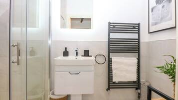Un baño moderno con cabina de ducha de vidrio, un lavabo blanco con espejo encima, un perchero negro y un cuadro en la pared.