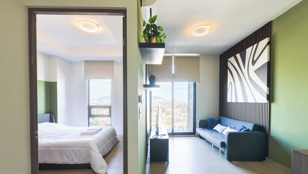 A modern hotel room with a bed, a sofa, a TV, and a large window with a city view. - Amo Living (Monterrey)