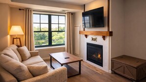Ein Wohnzimmer mit einer Couch, einem Couchtisch, einem Kamin, einem darüber montierten Fernseher und einem Fenster mit Vorhängen.