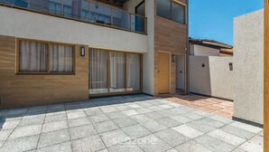 A modern two-story building with large windows, a balcony, and a tiled patio area. - Fully Equipped Aparts in Praia do Forte - PBS (Mata de São João)