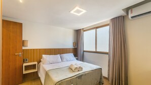 A bedroom with a bed, a bedside table, a wooden headboard, a window with curtains, and an air conditioning unit. - Fully Equipped Aparts in Praia do Forte - PBS (Mata de São João)