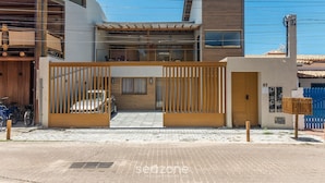 A modern two-story house with a wooden gate, a car parked inside, and a blue fence in the background. - Fully Equipped Aparts in Praia do Forte - PBS (Mata de São João)