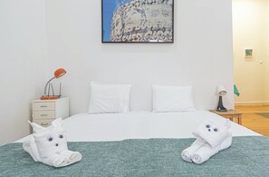 A hotel room with a bed, two white pillows, a green blanket, a nightstand with a lamp, and two white towels shaped like cats. - HOUZE Central Studio in Alcântara (Lisboa)