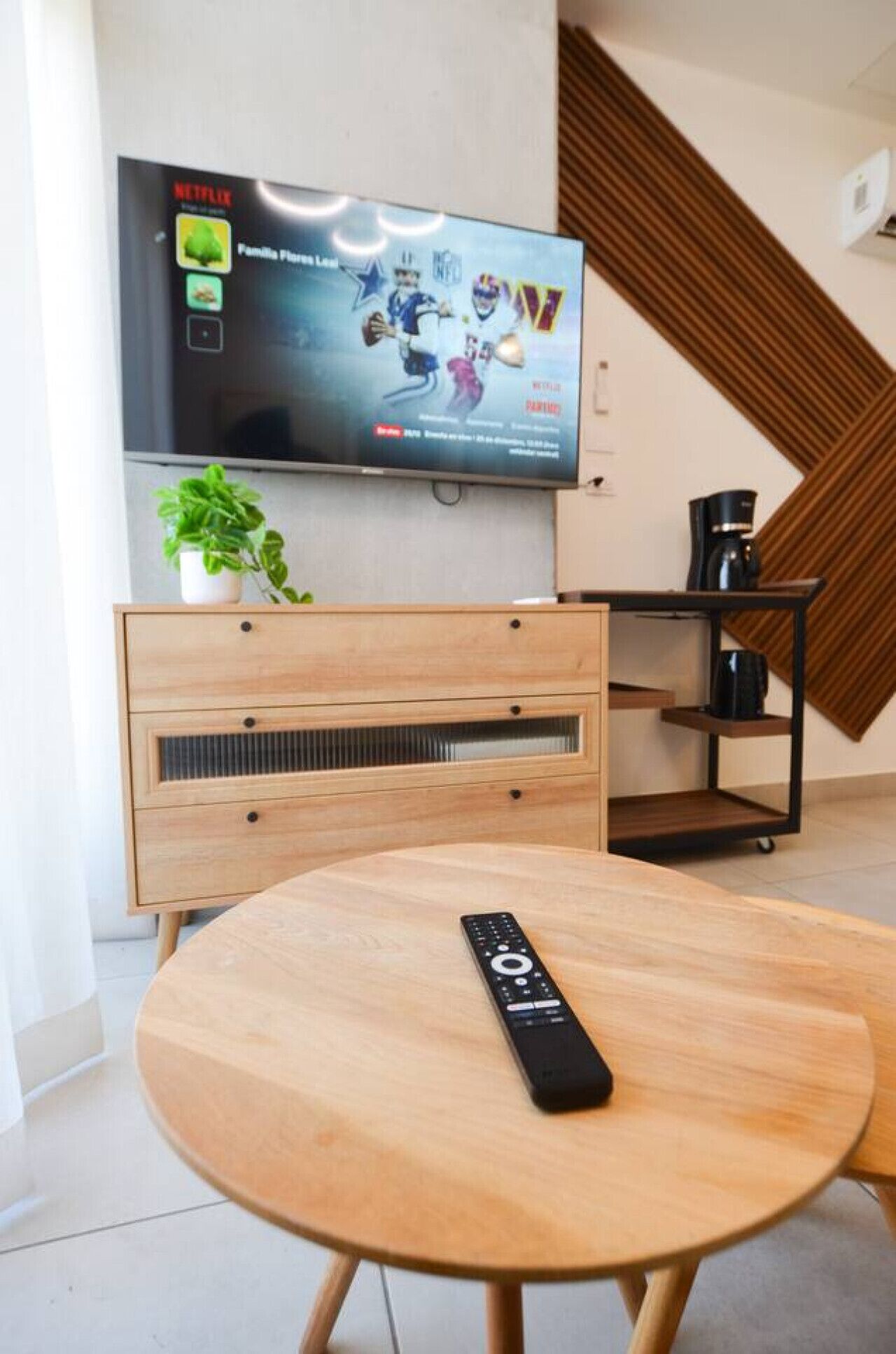 Un salon moderne avec un téléviseur fixé au mur, une table basse en bois, une télécommande, une plante et une machine à café.