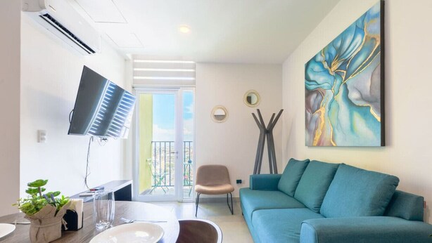 A modern living room with a teal sofa, a dining table with chairs, a TV mounted on the wall, and a balcony with outdoor furniture. - ¡Cerca de Fundidora! 6 ppl, GYM, Parking & 3 camas (Monterrey)