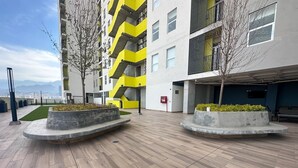 Un edificio moderno con fachada amarilla y gris, techo plano y un patio empedrado con jardineras y árboles.