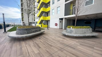 A modern building with a yellow and grey facade, a flat roof, and a paved courtyard with planters and trees.