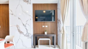 Una habitación moderna con pared de mármol, pared de paneles de madera, un televisor sobre un escritorio, una silla y un amplio ventanal con cortinas.