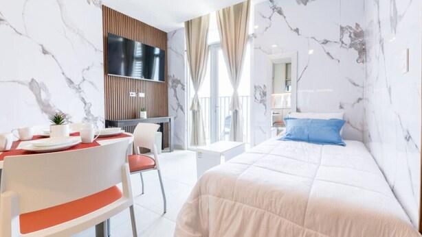 Una habitación de hotel moderna con pared de mármol, cama con almohada azul, zona de comedor con mesa roja y televisor montado en la pared.