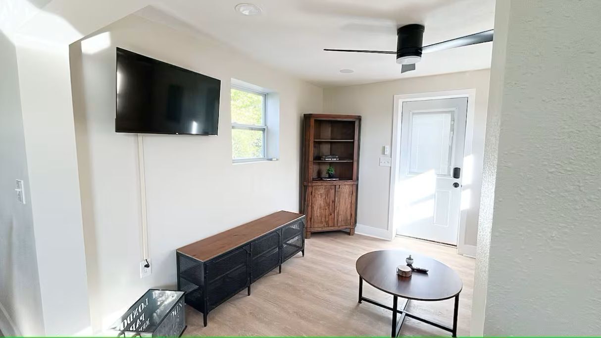 A modern living room with a flat-screen TV mounted on the wall, a wooden bench, a small round table, and a built-in wooden cabinet.