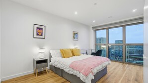 A modern bedroom with a large bed, a nightstand, a desk, and a window with a city view. - Entire City View Apartment 8 mins to London Brigde (London)