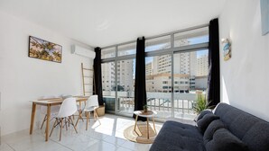 A modern living room with a sofa, dining table, chairs, a small side table, and a large window with curtains. - Studio Apartment 'Ocean - Pet Frendly' with Balcony, Wi-Fi and Air Conditioning (Portimão)