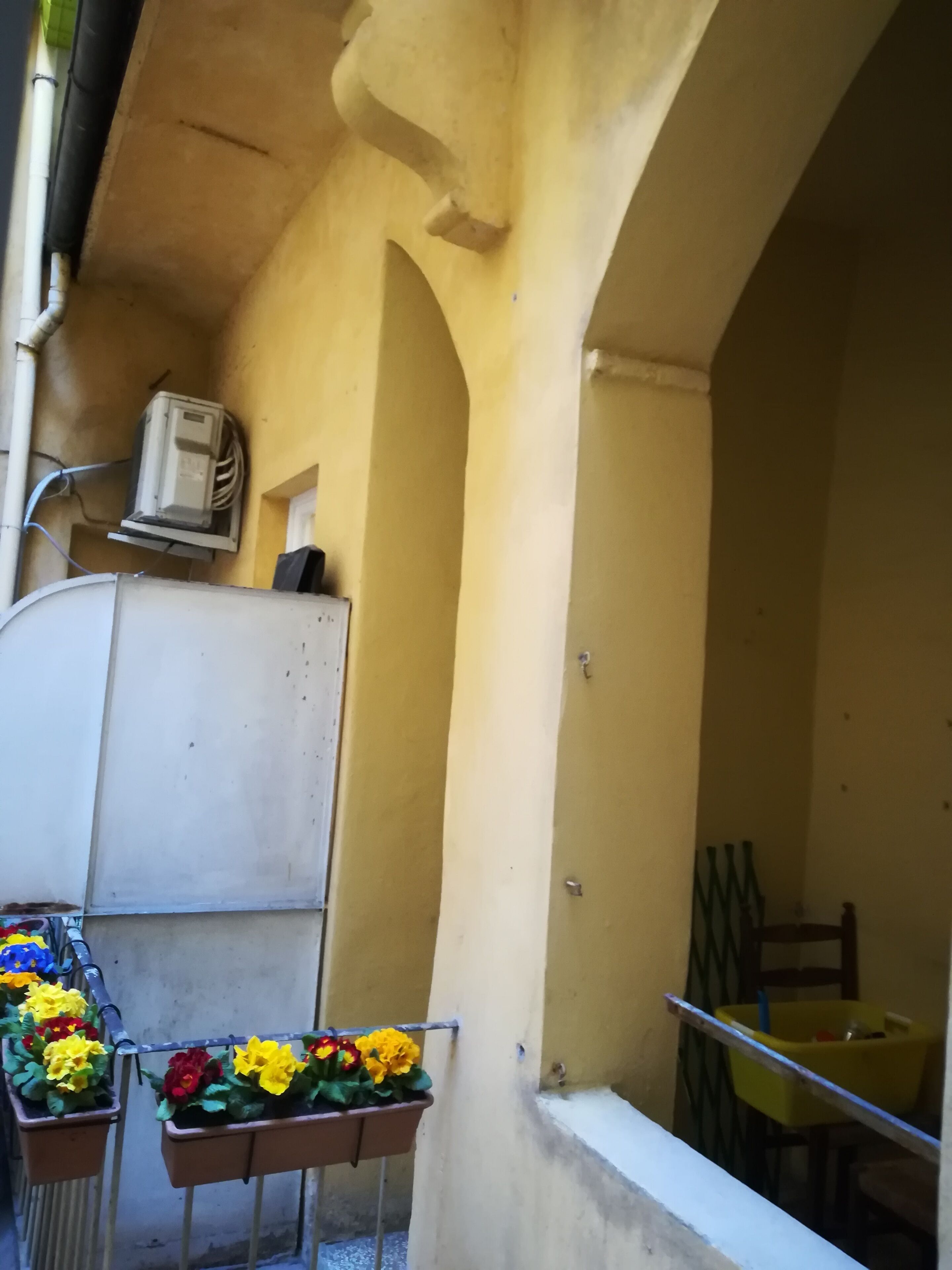 Ein Balkon mit einer beigefarbenen Wand, einer weißen Klimaanlage und Blumenkästen mit gelben und roten Blüten.