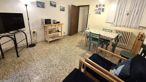 Ein kleiner, möblierter Raum mit Fernseher, Mikrowelle, Tisch mit Stühlen, Heizkörper und einer Tür zu einem anderen Raum.