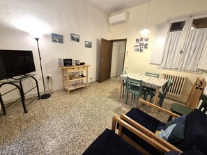 Ein kleiner, möblierter Raum mit Fernseher, Mikrowelle, Tisch mit Stühlen, Heizkörper und einer Tür zu einem anderen Raum.