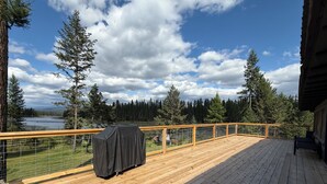 Um deck de madeira com churrasqueira coberta, um banco e vista para um lago e floresta.