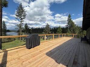 Een houten terras met een overdekte barbecue, een bank en uitzicht op een meer en bos.