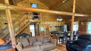 Interior rústico de cabana com escada de madeira, sofá, mesa de jantar e poltrona de couro.